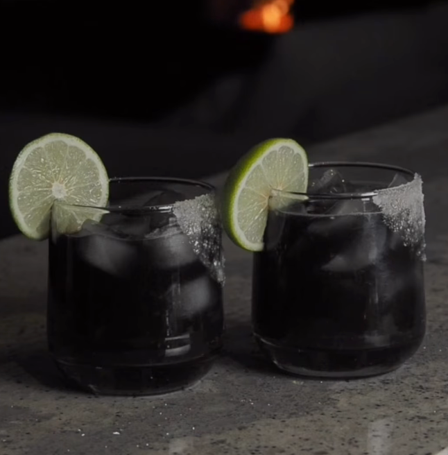 Two black margaritas garnished with salt rim, hot honey & lime wedge.